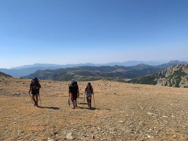 Trois randonneurs marchent sur un terrain rocailleux avec des montagnes en arrière-plan.
