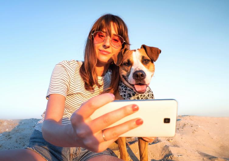 Une jeune femme et son chien sourient en prenant un selfie sur la plage.