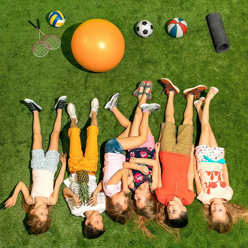 Une vue aérienne dun groupe denfants allongés sur lherbe avec un ballon de basket, un ballon de football, une raquette de badminton et un ballon de volley-ball.