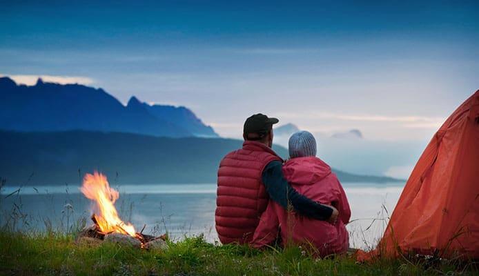 Un couple regarde un feu de camp devant un lac et des montagnes.