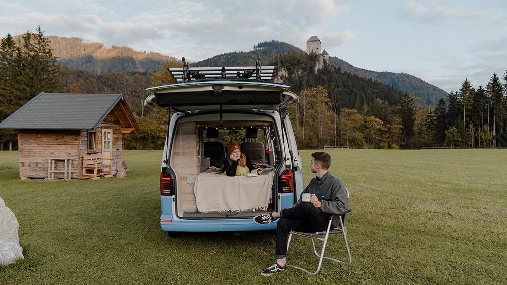 Un couple est assis devant leur van bleu et blanc, en train de profiter de la vue sur les montagnes.