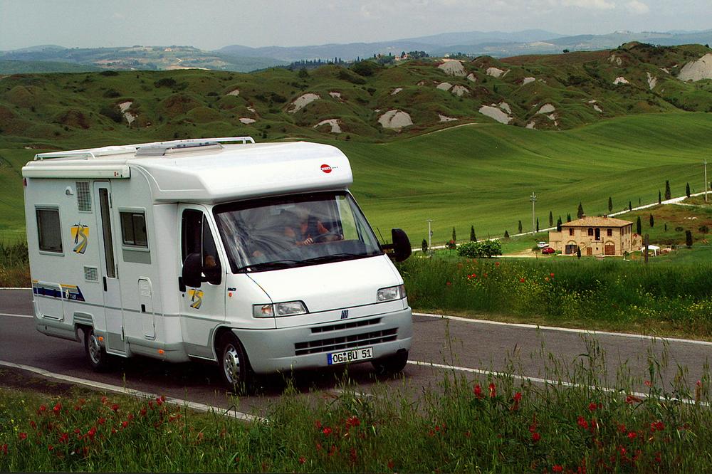 Un camping-car blanc roule sur une route de campagne, avec des collines verdoyantes en arrière-plan.