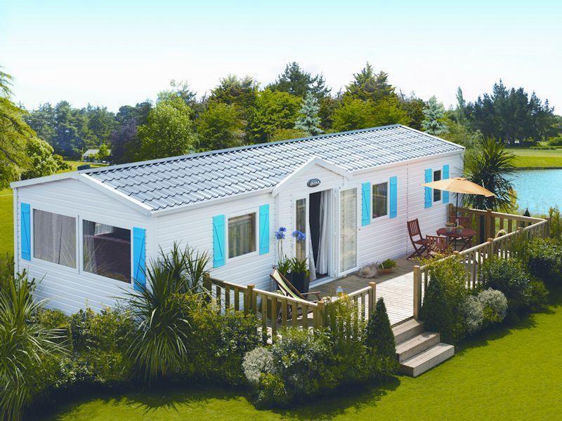 Une maison mobile blanche avec des volets bleus est située dans un camping près dun lac.