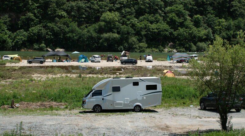 Une aire de camping avec des tentes, des caravanes et des voitures garées à côté dune rivière.