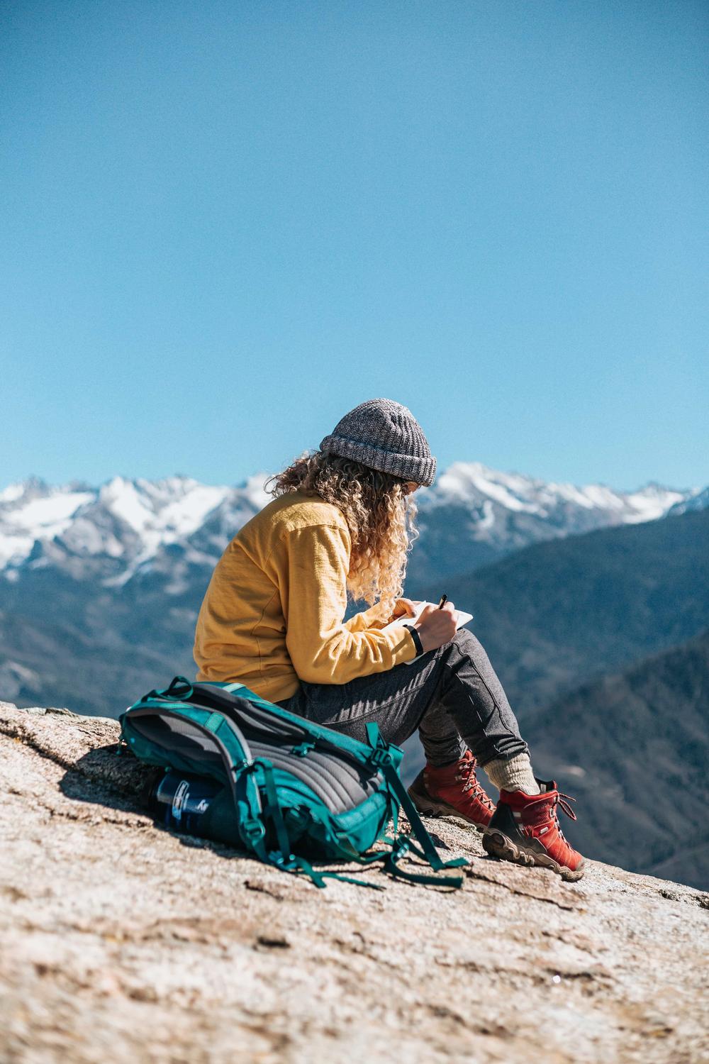 Une femme est assise sur un rocher et écrit dans son carnet avec une montagne enneigée en arrière-plan.