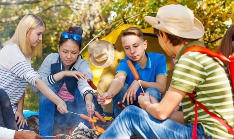 Des adolescents sont assis autour dun feu de camp et font griller des saucisses.