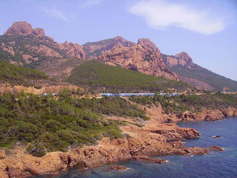 Un train bleu et blanc longeant la côte rocheuse de la mer Méditerranée avec des montagnes en arrière-plan.