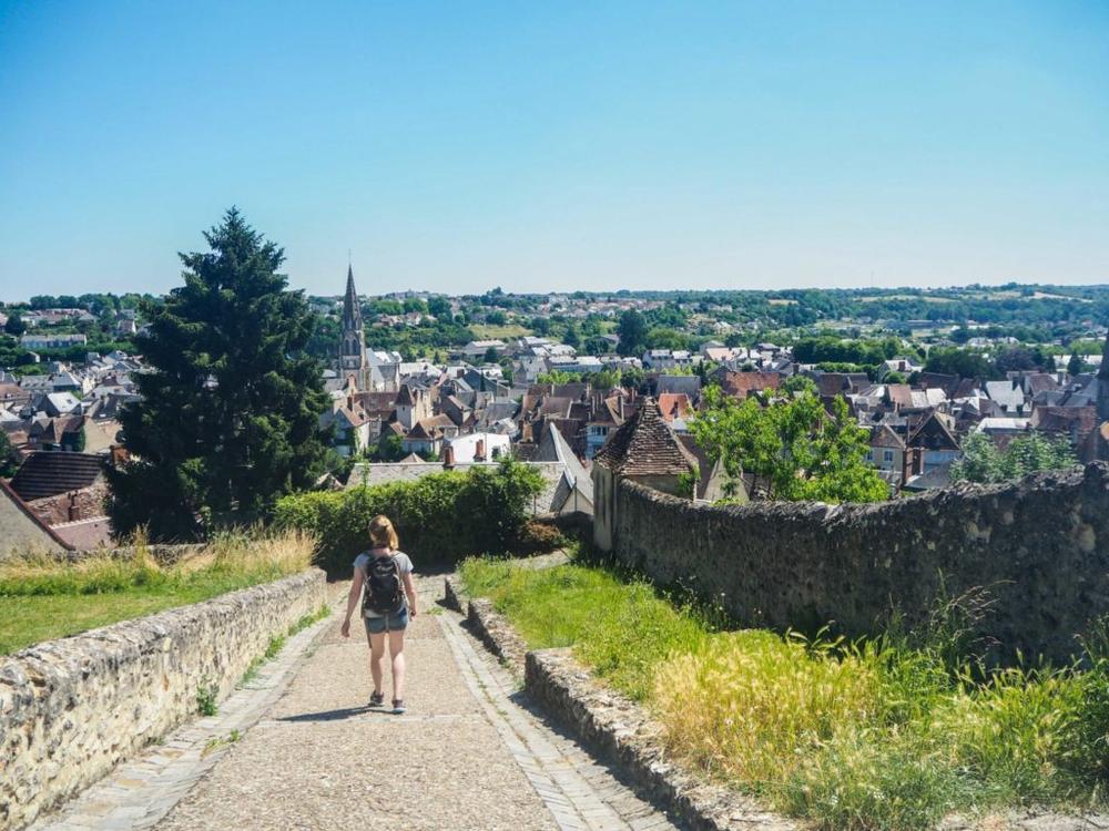 Une femme marche sur un chemin en pierre qui descend vers une ville médiévale française.
