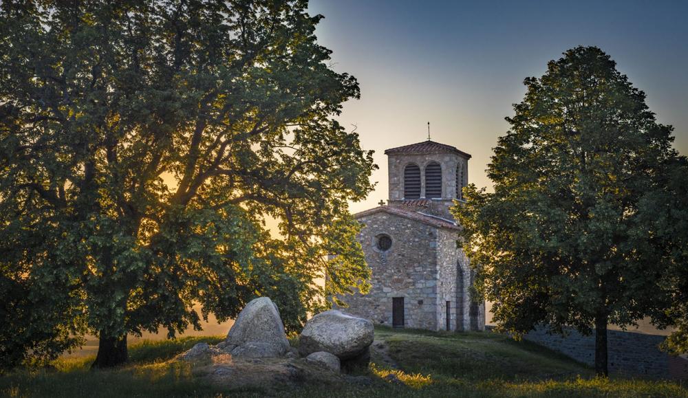 Une église en pierre se dresse sur une colline, entourée darbres et de verdure.