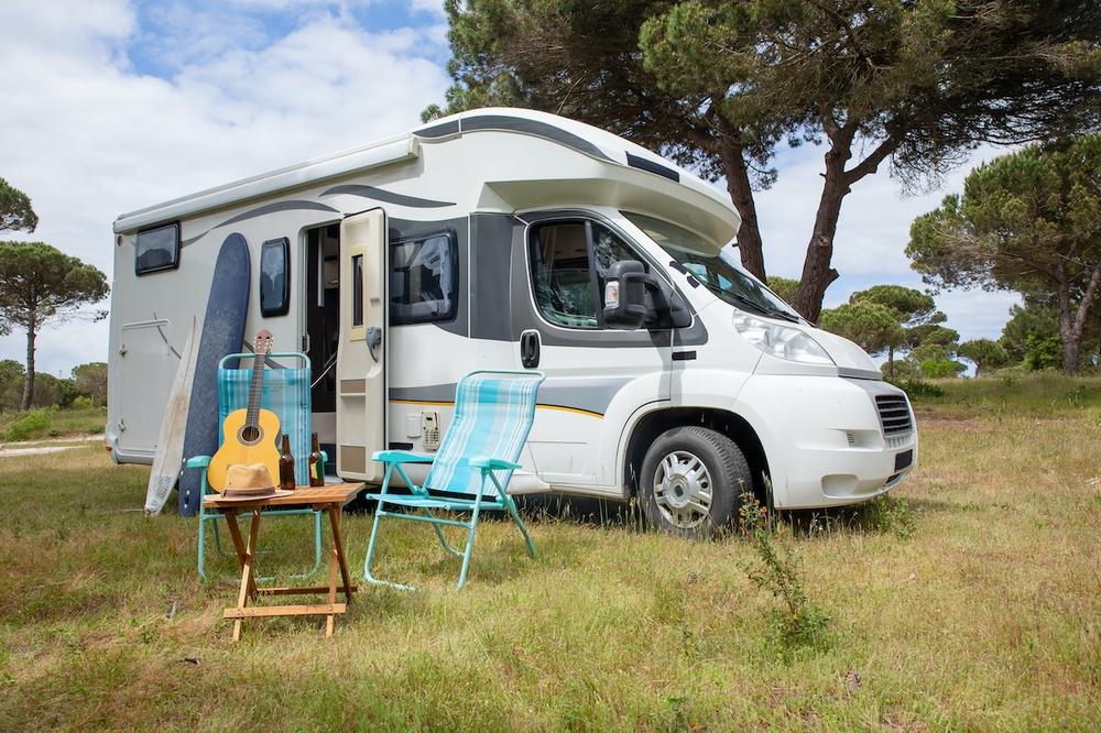 Une image dun camping-car garé dans un camping avec des chaises de camping, une table, une guitare et une planche de surf à lextérieur.