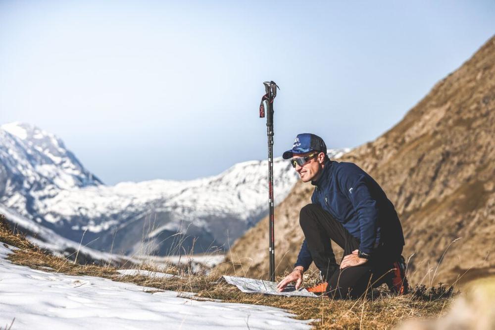 Un homme accroupi consulte une carte sur un fond de montagne enneigée.
