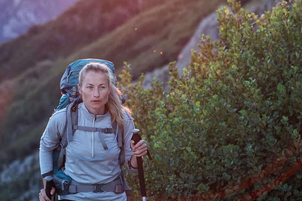 Une femme blonde en vêtements de randonnée et sac à dos marche sur un sentier en montagne.