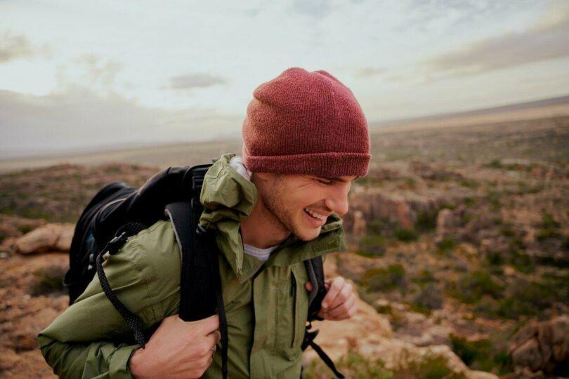 Un jeune homme portant un sac à dos et un bonnet sourit en marchant sur un sentier de montagne.