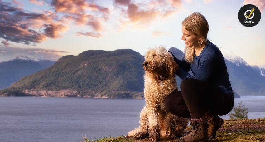 Une femme accroupie à côté de son chien sur un promontoire rocheux, avec un lac et des montagnes en arrière-plan.