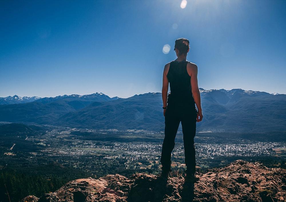 Une femme est debout sur un rocher et regarde au loin une ville et des montagnes.