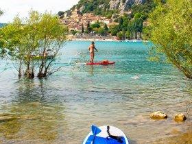 Une planche de paddle bleue est posée sur le rivage avec en fond un homme pagayant sur un lac entouré darbres et de montagnes.