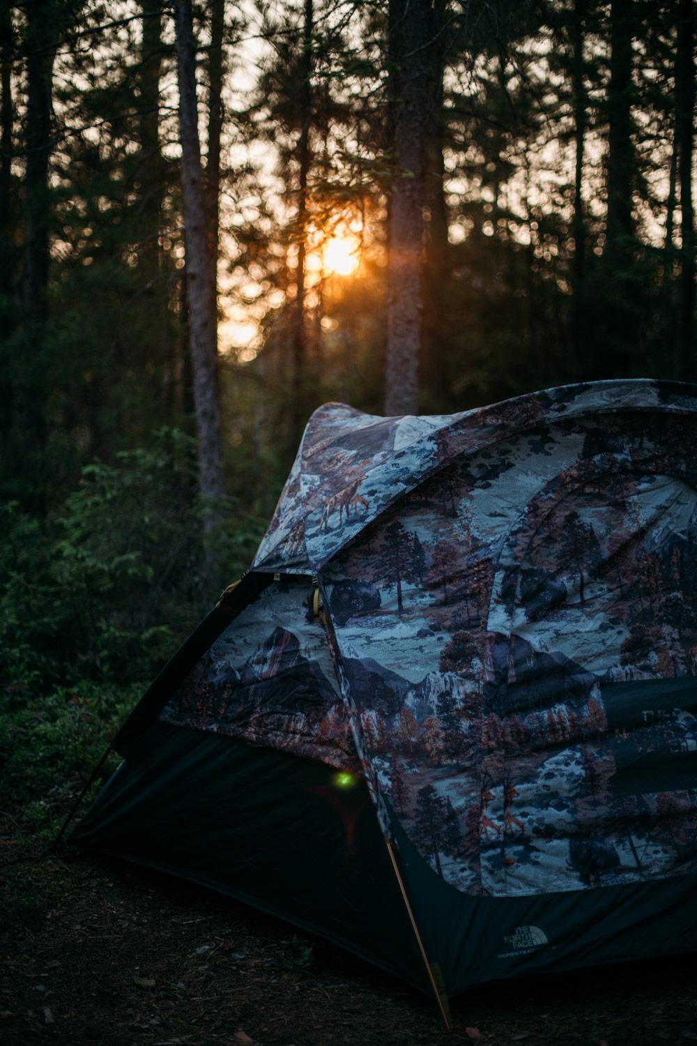 Une tente de camping avec un motif camouflage est installée dans une forêt au coucher du soleil.