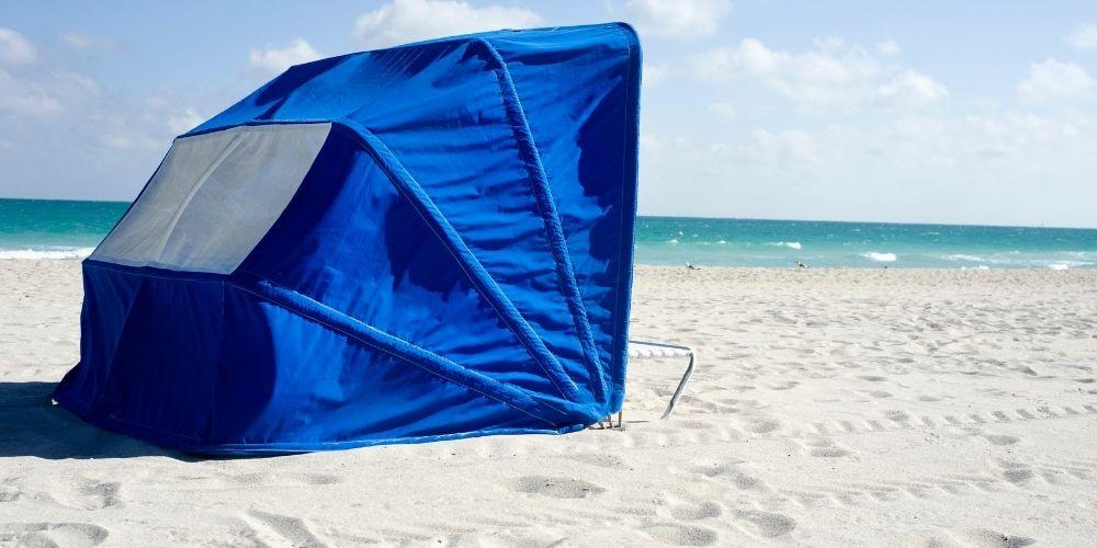Une tente de plage bleue est installée sur le sable avec la mer en fond.