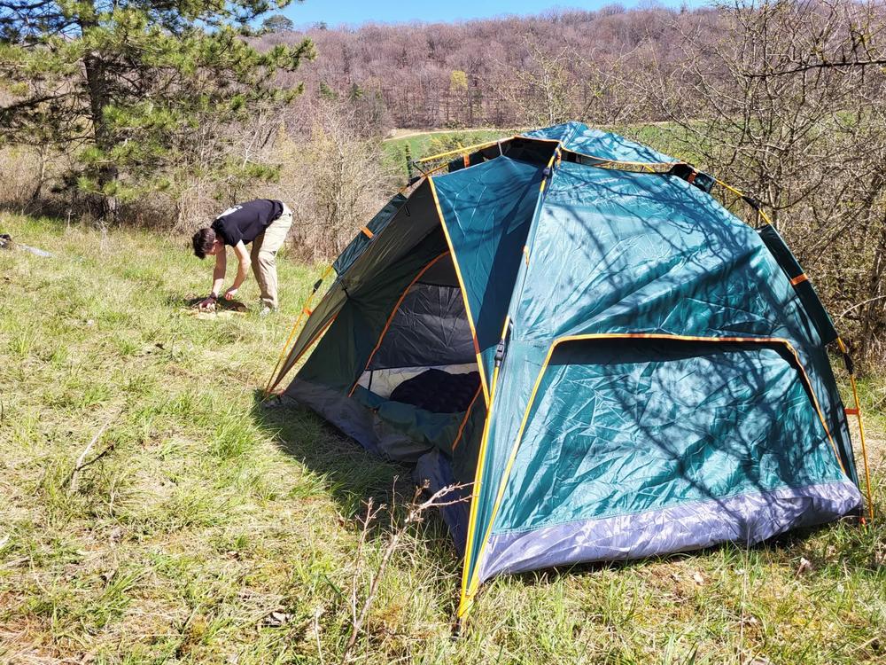 Un homme installe une tente verte et grise dans une clairière.