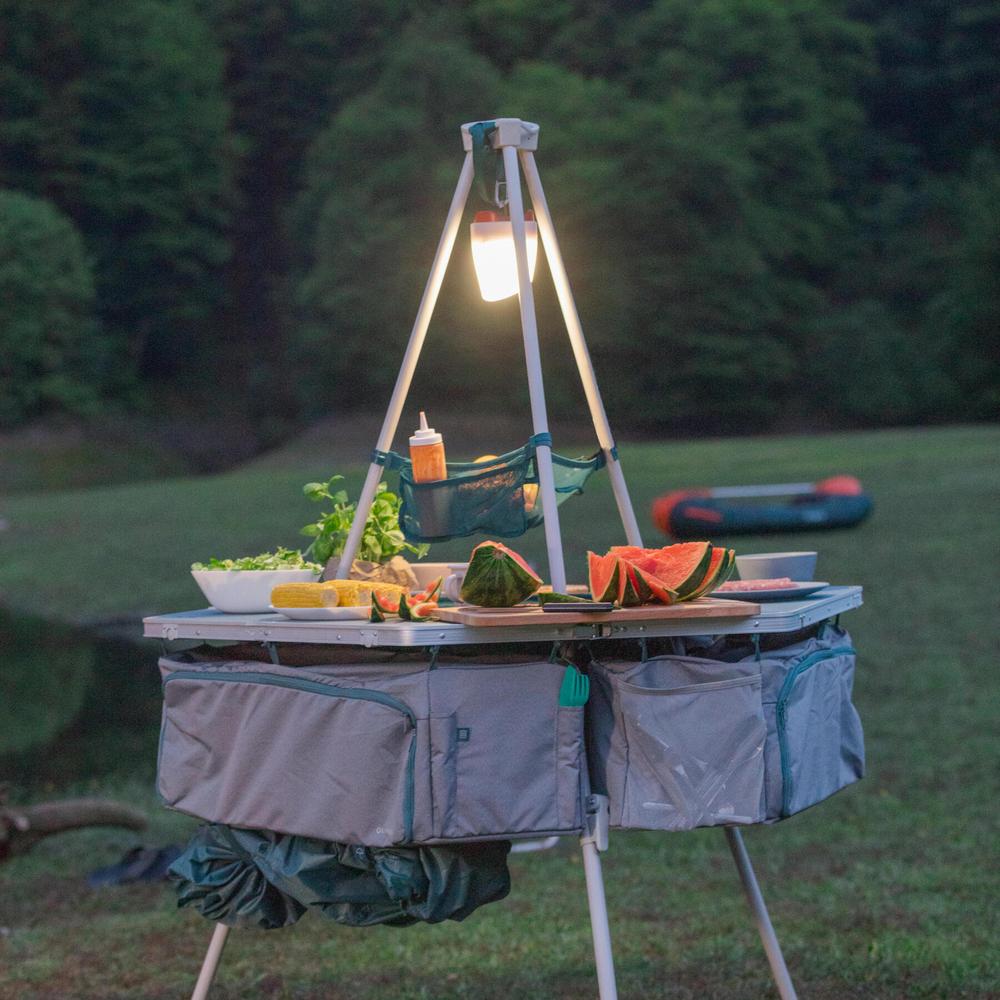 Une table de camping pliable avec un dessus en aluminium et des pieds en acier, avec une lanterne suspendue au-dessus.