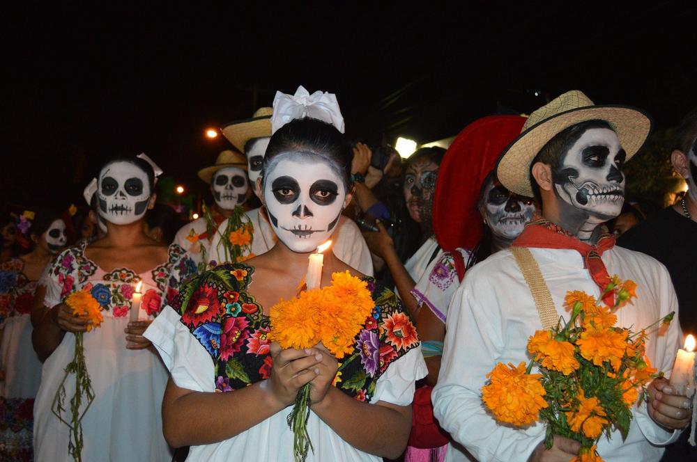 Des personnes portant des costumes et des maquillages de squelettes, défilent en tenant des bougies et des fleurs.