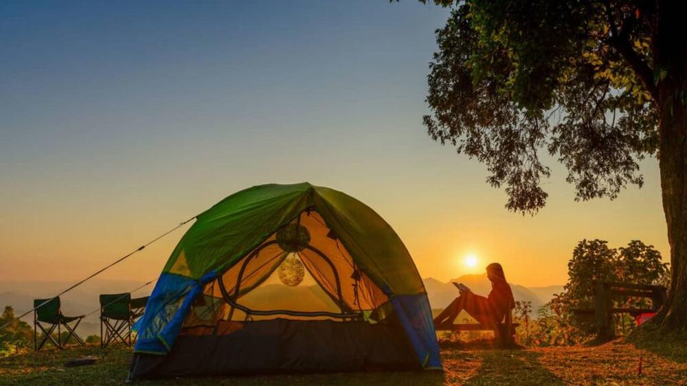 Une femme est assise sur une chaise de camping et lit un livre devant une tente verte au coucher du soleil.