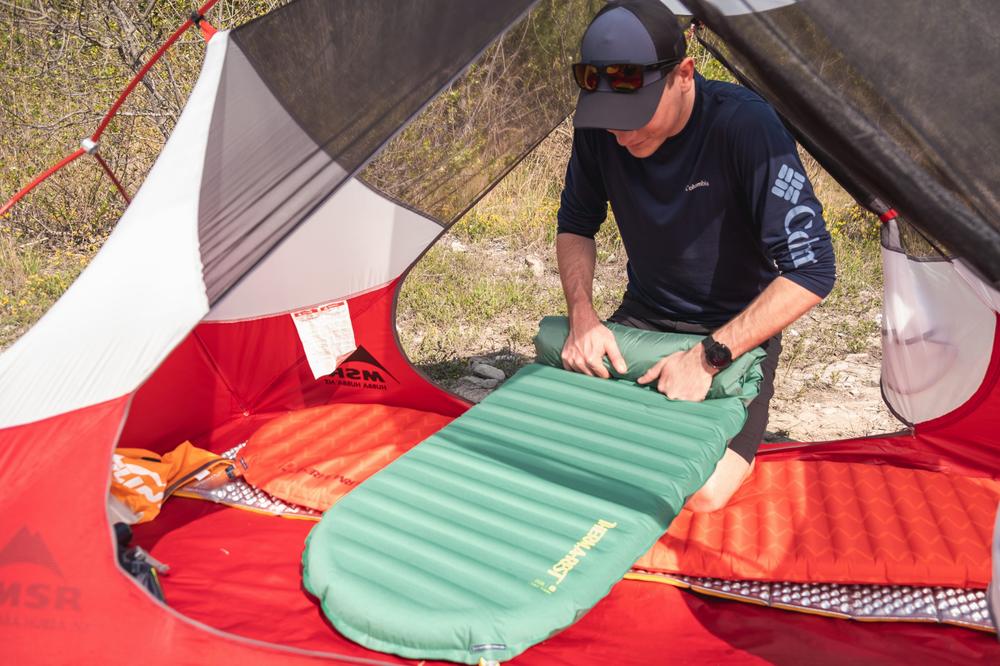 Un homme ajuste un matelas de camping vert dans une tente.