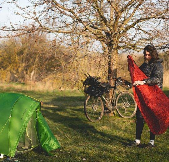 Un homme est en train de plier une bâche rouge devant une tente verte et un vélo.