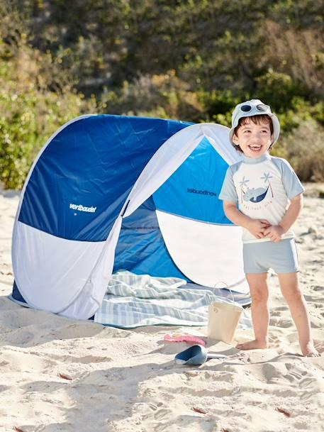 Un petit garçon souriant se tient à côté dune tente de plage bleue et blanche.