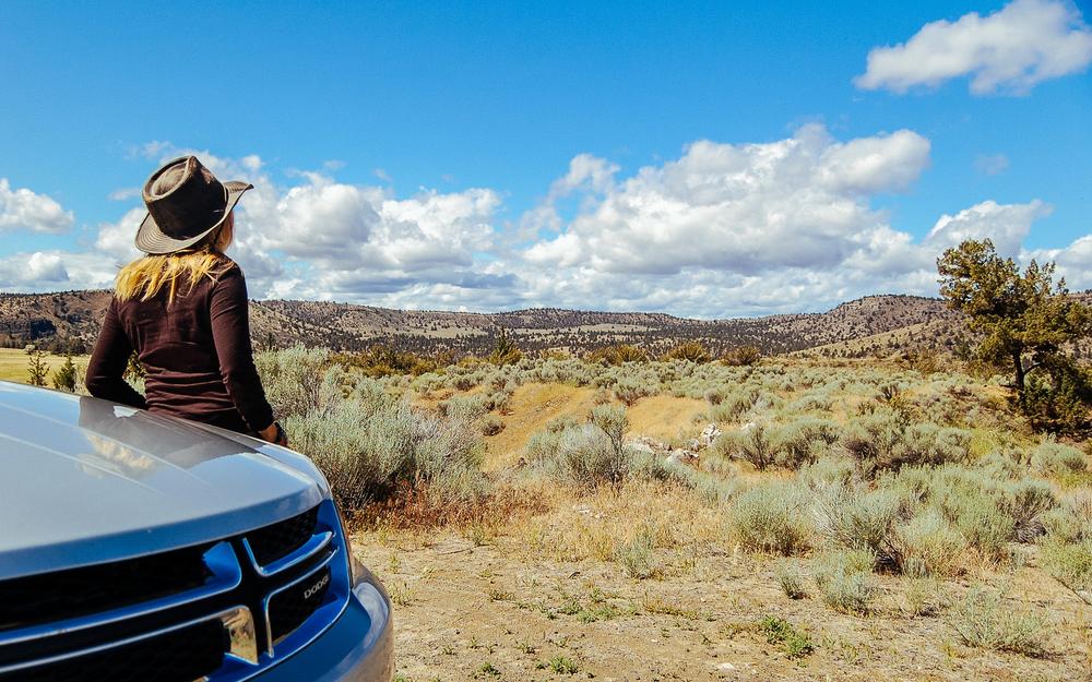 Une femme portant un chapeau de cowboy est accoudée sur le capot dune voiture et regarde le paysage.