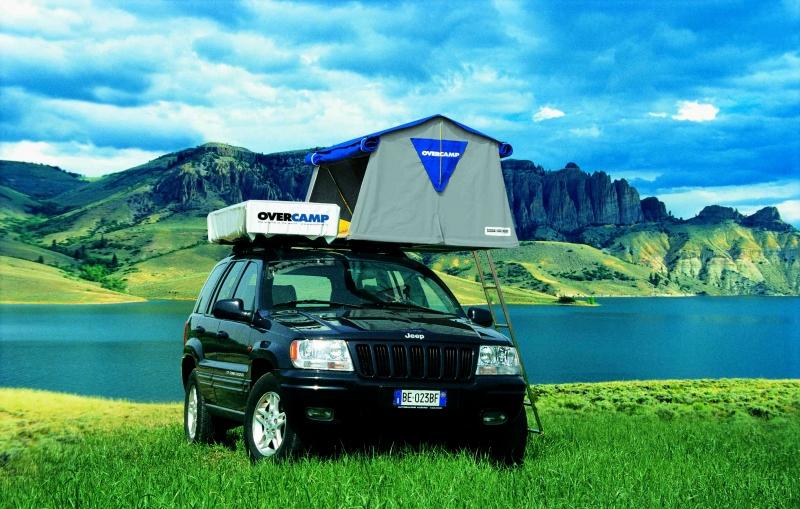 Une Jeep noire avec une tente sur le toit est garée dans un champ avec un lac et des montagnes en arrière-plan.